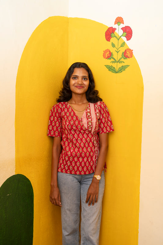 A woman stands in front of a yellow arched backdrop with painted flowers, wearing a red patterned short-sleeve blouse and light gray jeans.