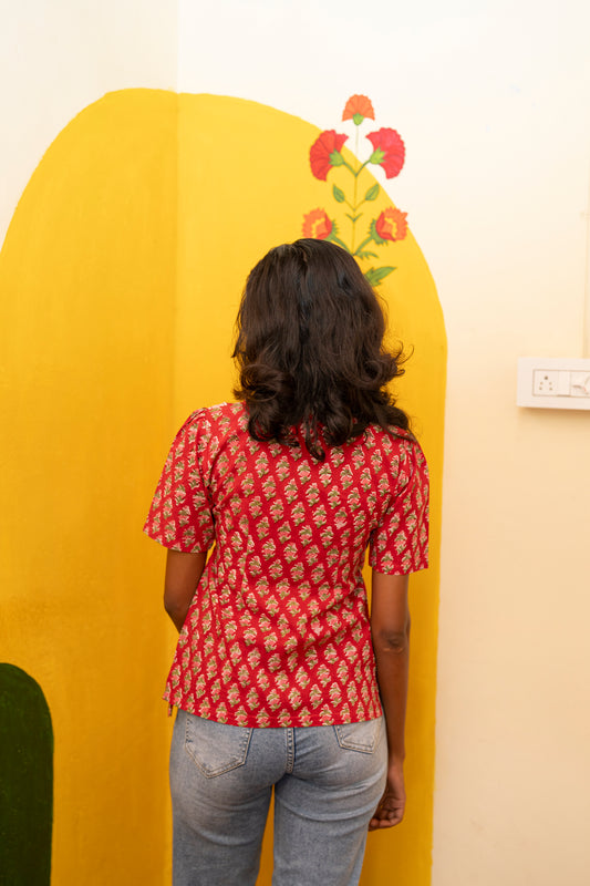 A woman viewed from behind wearing a red short-sleeve t-shirt with a white geometric block print pattern and light blue jeans, standing in front of a yellow decorative wall panel.