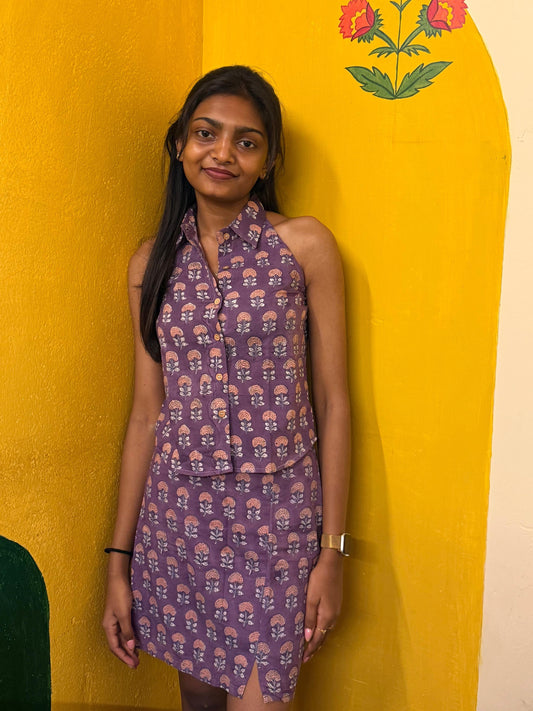 Woman in a purple patterned dress standing against a yellow wall with floral designs.