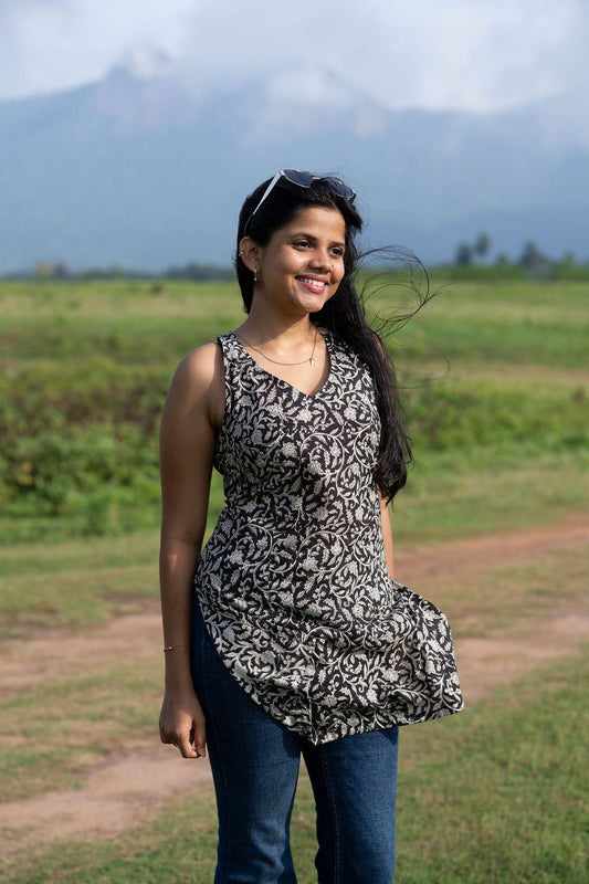 A woman smiling outdoors in a sleeveless black and white patterned top with blue jeans, standing in a grassy field with mountains in the background.