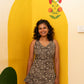 A woman with curly dark hair stands smiling in front of a yellow arched wall backdrop. She is wearing a sleeveless black and beige patterned peplum top and light beige pants.