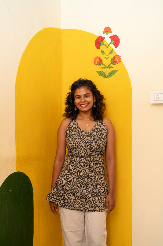 A woman with curly dark hair stands smiling in front of a yellow arched wall backdrop. She is wearing a sleeveless black and beige patterned peplum top and light beige pants.