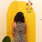 A woman with curly dark hair stands with her back to the camera, wearing a sleeveless black and white patterned top and white pants, posing in front of a bright yellow painted wall corner decorated with a small flower motif.
