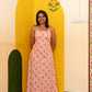A woman stands barefoot in front of a painted yellow wall corner, wearing a sleeveless pink maxi dress with small repeating motifs.