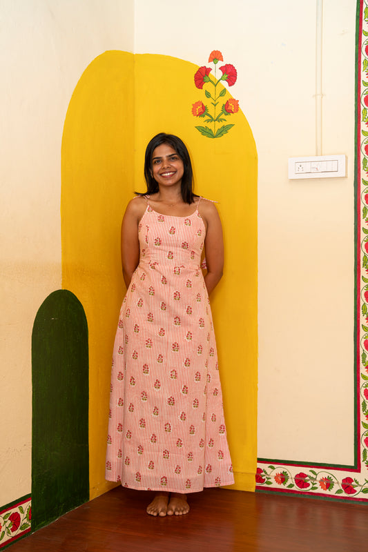 A woman stands barefoot in front of a painted yellow wall corner, wearing a sleeveless pink maxi dress with small repeating motifs.
