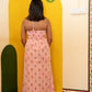 A woman stands with her back to the camera wearing a pink sleeveless maxi dress with a small red and green block print pattern. She is positioned in front of a large yellow archway prop in an indoor setting with a wooden floor.