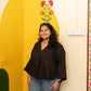 A woman with long dark hair stands smiling in front of a cream-colored wall decorated with a large yellow arch, green leaf shapes, and a red and orange floral motif. She is wearing a dark brown V-neck blouse with wide three-quarter sleeves and blue jeans.