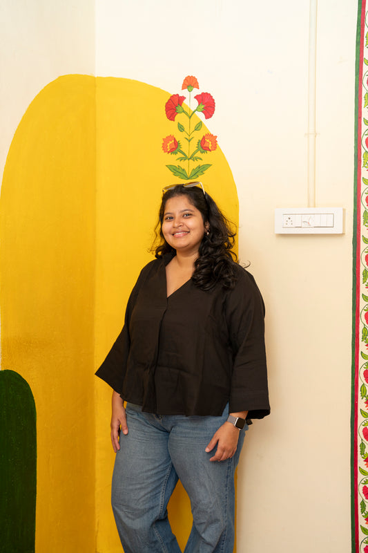 A woman with long dark hair stands smiling in front of a cream-colored wall decorated with a large yellow arch, green leaf shapes, and a red and orange floral motif. She is wearing a dark brown V-neck blouse with wide three-quarter sleeves and blue jeans.