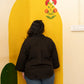 A person viewed from behind wearing a dark brown jacket and blue jeans, standing in front of a large yellow decorative wall panel with painted flowers and a green shape against a cream-colored wall.