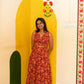 A woman wearing a sleeveless red dress with a large orange floral pattern and a tied waist detail, standing in front of a bright yellow wall with painted floral motifs.