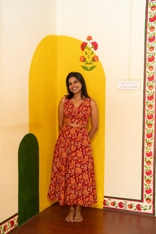 A woman wearing a sleeveless red dress with a large orange floral pattern and a tied waist detail, standing in front of a bright yellow wall with painted floral motifs.