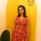 A woman wearing a sleeveless red dress with a large orange floral pattern and a tied waist detail, standing in front of a bright yellow wall with painted floral motifs.
