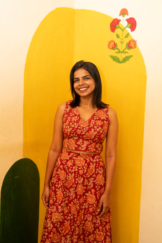 A woman wearing a sleeveless red dress with a large orange floral pattern and a tied waist detail, standing in front of a bright yellow wall with painted floral motifs.