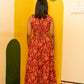 A woman viewed from behind wearing a sleeveless red dress with a large orange floral pattern and a tied waist detail, standing in front of a bright yellow wall with painted floral motifs.