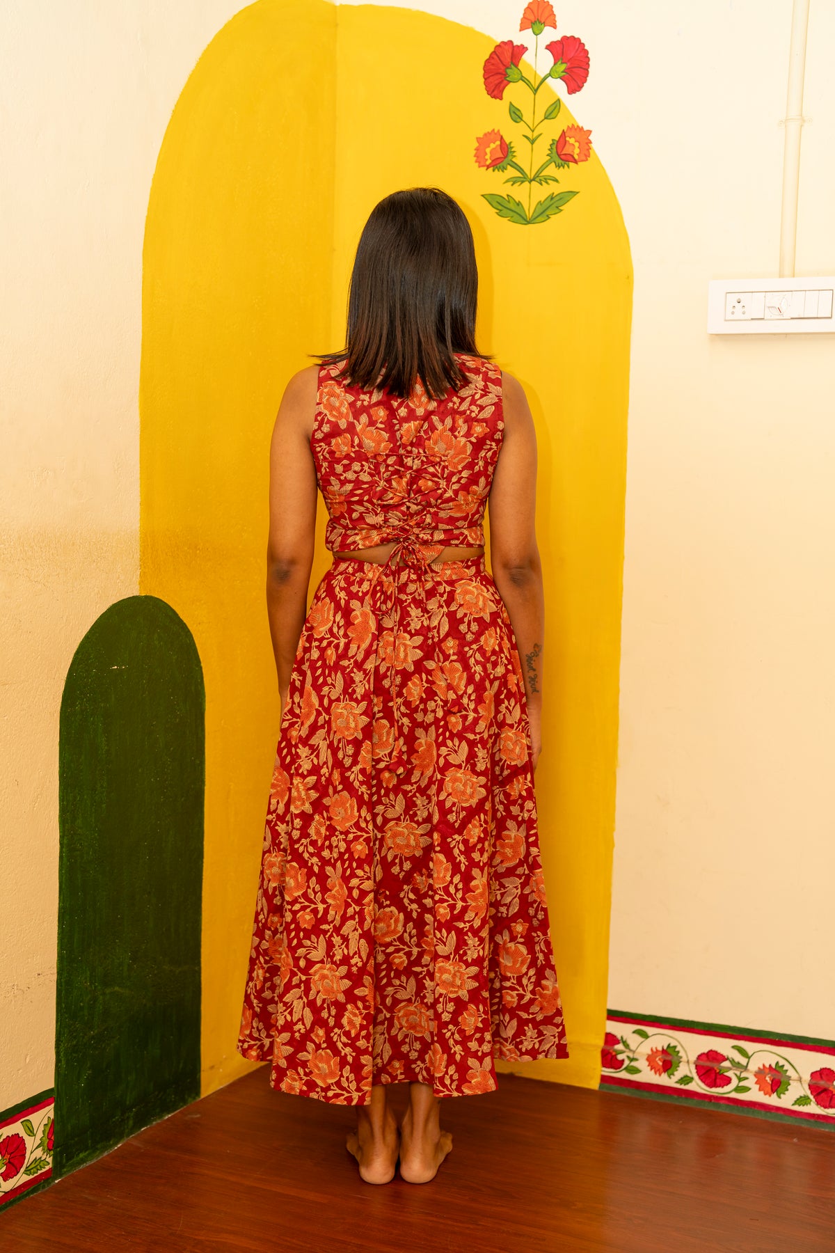 A woman viewed from behind wearing a sleeveless red dress with a large orange floral pattern and a tied waist detail, standing in front of a bright yellow wall with painted floral motifs.