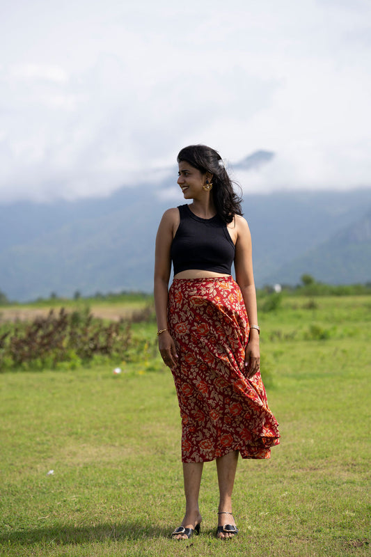 A woman stands in a grassy field with mountains and a cloudy sky in the background, wearing a black sleeveless crop top and a red floral midi skirt.