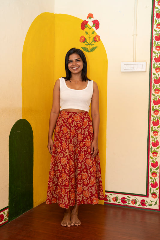 A woman stands barefoot in front of a painted yellow and cream wall, wearing a white sleeveless crop top and a long red-orange floral skirt.