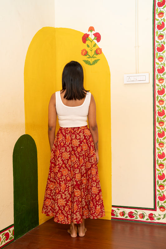 A woman viewed from behind wearing a sleeveless white top and a red floral maxi skirt, standing in a room with a yellow painted arch on the wall.