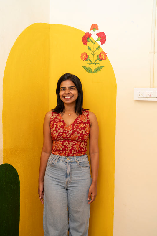 A woman smiling and posing in front of a yellow arched wall backdrop. She is wearing a sleeveless red and orange floral print top paired with light-wash blue jeans.