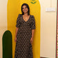 A woman stands indoors wearing a long black dress with a beige floral print. She is positioned in front of a decorative yellow and cream wall with painted arch shapes and floral details.