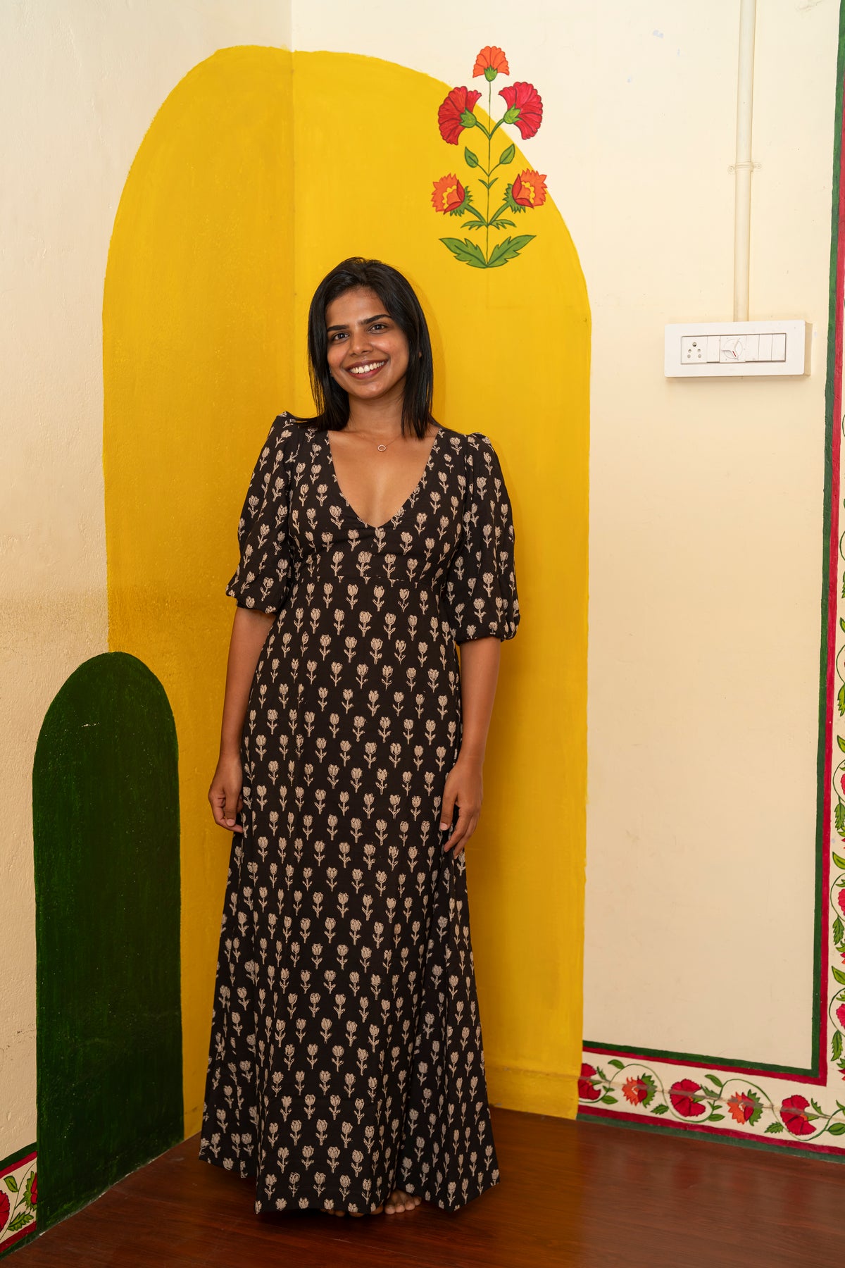 A woman stands indoors wearing a long black dress with a beige floral print. She is positioned in front of a decorative yellow and cream wall with painted arch shapes and floral details.