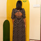 A woman viewed from behind wearing a black maxi dress with a beige floral pattern and a open back, standing in front of a yellow painted wall corner.