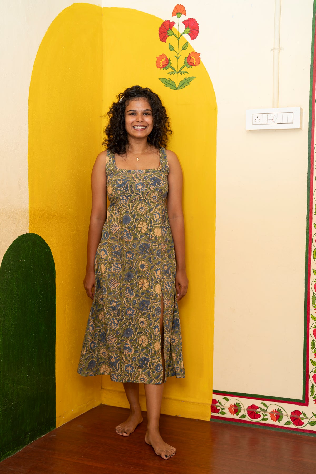 A woman stands barefoot in a colorful room wearing a sleeveless midi dress with a blue and gold floral block print pattern.