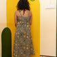 A woman viewed from behind wearing a sleeveless midi dress with a blue, green, and beige floral pattern, standing in front of a yellow painted wall corner.