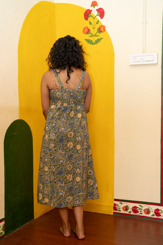 A woman viewed from behind wearing a sleeveless midi dress with a blue, green, and beige floral pattern, standing in front of a yellow painted wall corner.