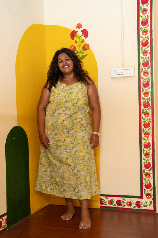 A woman stands barefoot in a room corner wearing a sleeveless, ankle-length yellow-green floral dress. The background features a painted yellow arch, a green arch, and a decorative red and green border on the cream-colored wall.