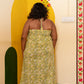 A woman viewed from behind wearing a strapless halter midi dress with a yellow and green floral pattern, standing in a room with a yellow painted arch on the wall.