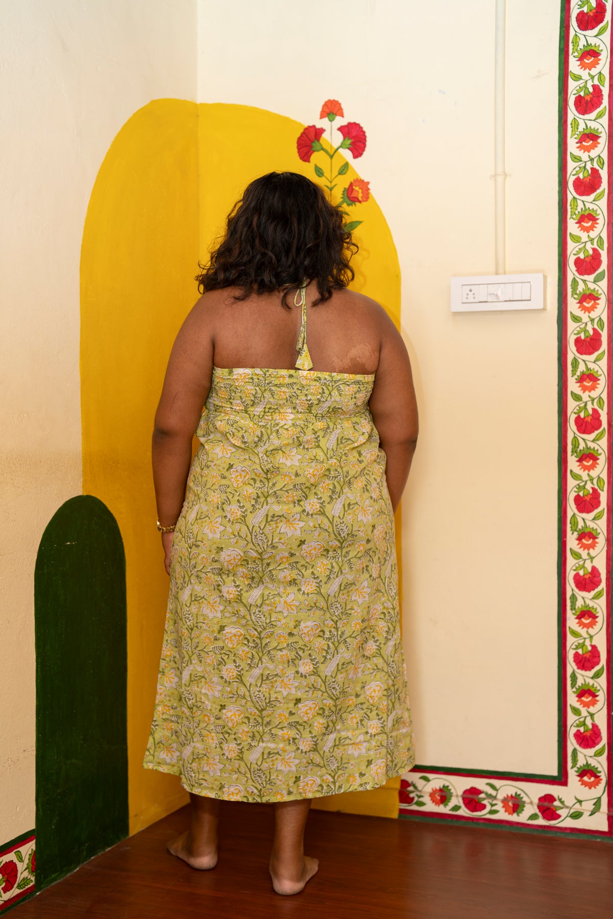 A woman viewed from behind wearing a strapless halter midi dress with a yellow and green floral pattern, standing in a room with a yellow painted arch on the wall.